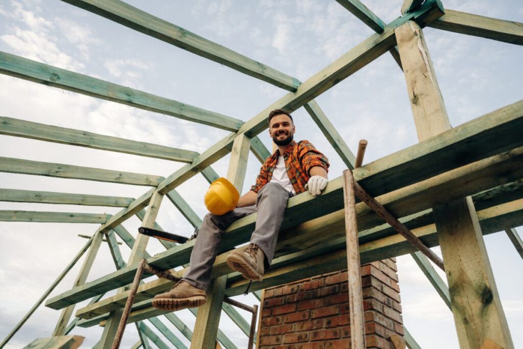 Un ouvrier de bâtiment souriant assis sur la charpente de la maison